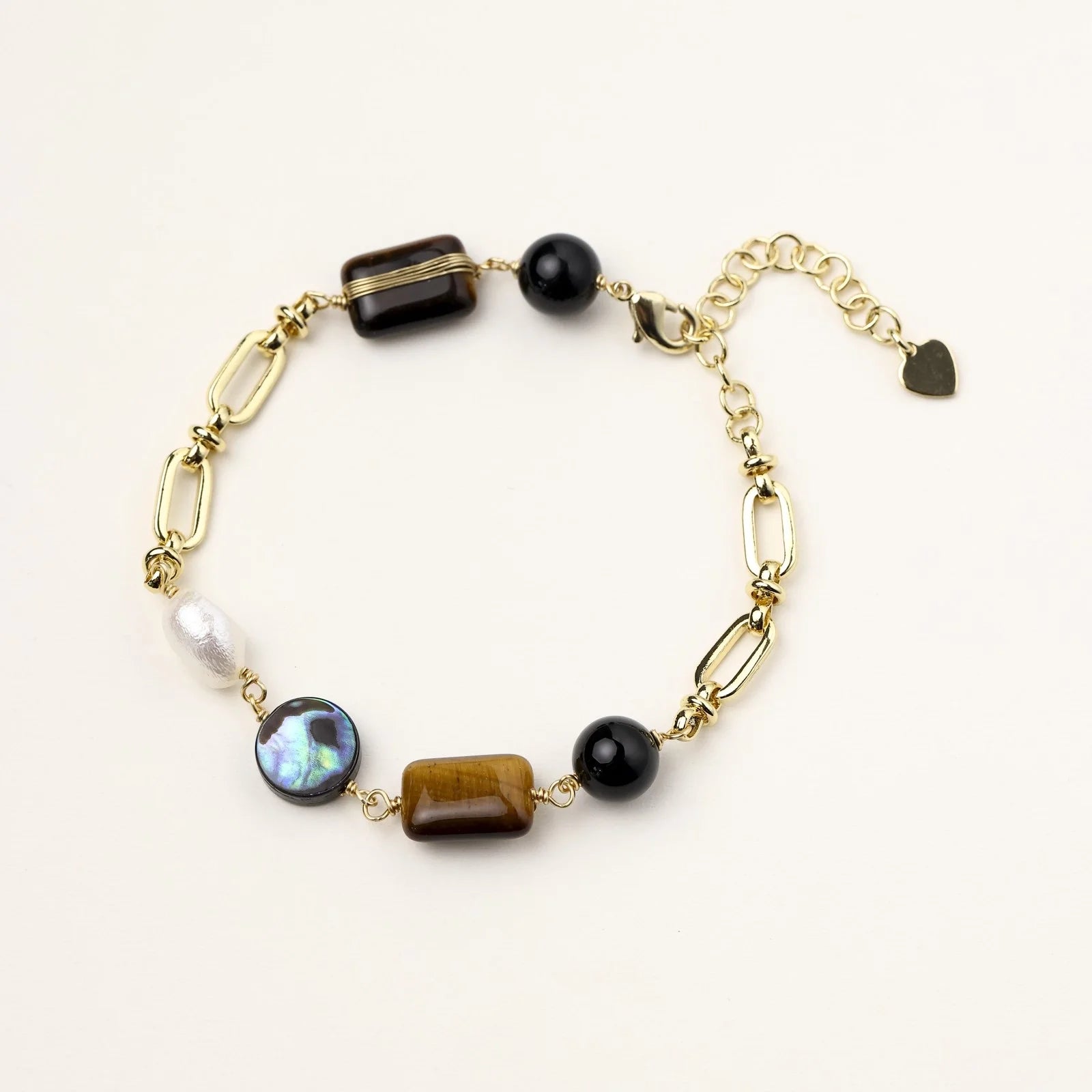 Gold chain bracelet with tiger's eye, obsidian, pearl, and iridescent abalone stones on cream background.