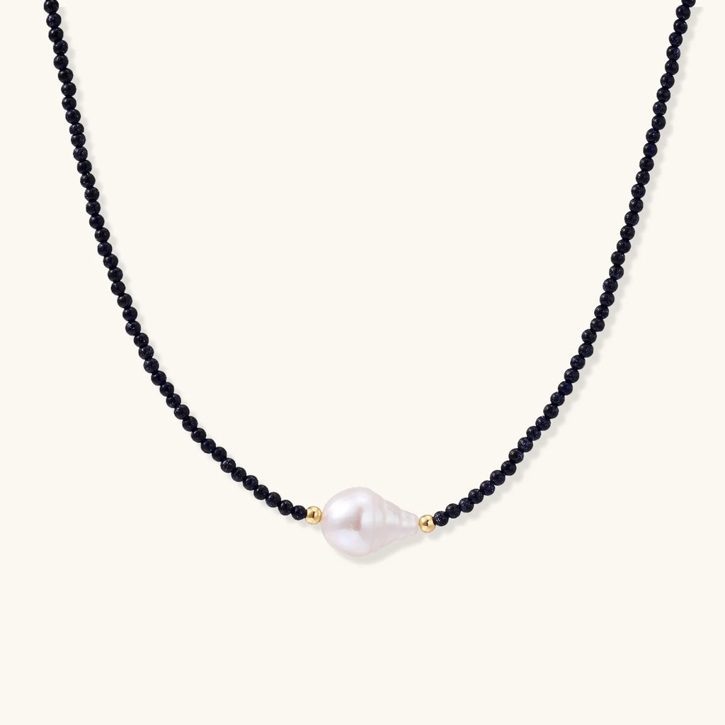 Delicate necklace with black blue sandstone beads and baroque pearl center on white background with gold accents.