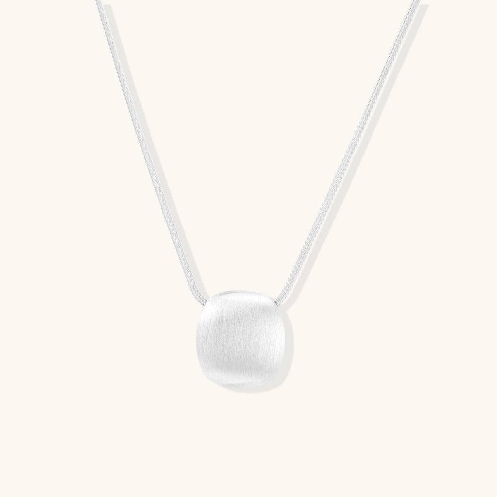 Minimalist brushed sterling silver dome pendant on delicate chain against cream background, showing matte textured finish