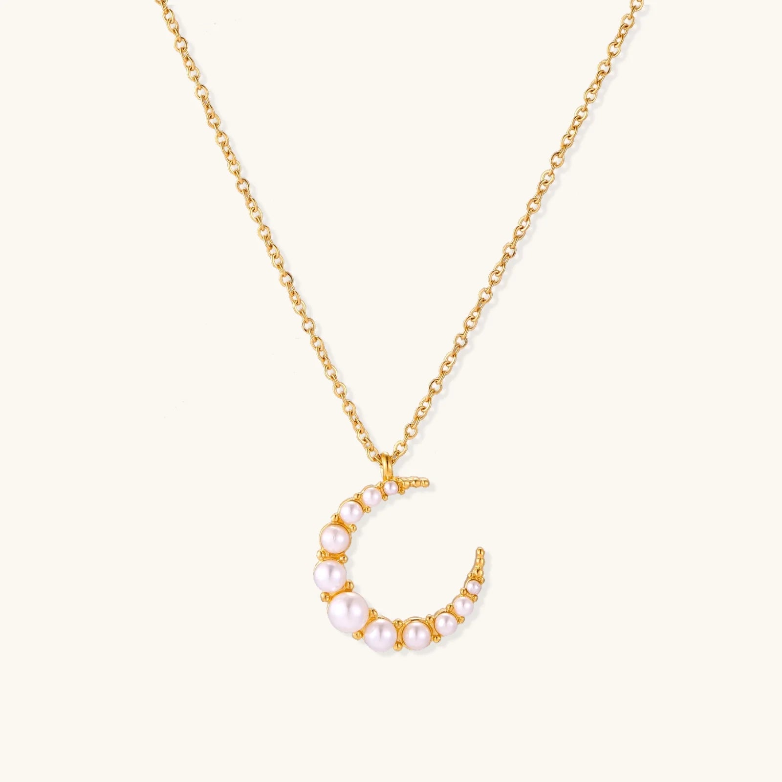 Gold crescent moon pendant with graduated white pearls on delicate chain against cream background